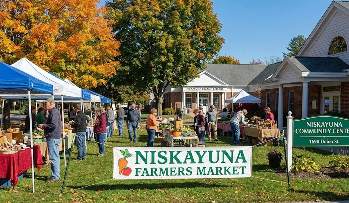 An outdoor scene of the Niskayuna Farmers Market with people browsing vendors under white and blue tents on a sunny day. A banner in the foreground reads "NISKAYUNA FARMERS MARKET," and a sign to the right marks the "Niskayuna Community Center at 1690 Union St.